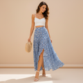 A woman stands outdoors in a sunlit courtyard wearing a white spaghetti-strap crop top and a flowing blue floral high-low maxi skirt, holding a woven straw bag.