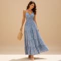 A woman wearing a light blue floral tiered maxi dress stands outdoors in a Mediterranean-style courtyard with white walls, blue doors, and purple flowers. She is holding a woven straw handbag and lifting the skirt slightly to show its flow.