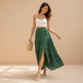 A woman wearing a white spaghetti-strap crop top and a high-waisted, flowing green sequined maxi skirt with a front slit, walking outdoors in a sunlit courtyard with potted plants and a wooden door in the background.