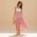 A woman stands outdoors on a sunlit stone walkway wearing a white cropped tank top and a flowing pink floral maxi skirt, holding a woven handbag. White buildings, potted plants, and pink flowers are visible in the background.