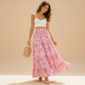 A woman stands outdoors in a sunlit courtyard wearing a white spaghetti-strap crop top and a long pink floral maxi skirt, holding out the skirt to show its flow. She carries a woven straw tote and wears beige sandals.