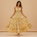 A woman wearing a yellow floral spaghetti-strap maxi dress with a tiered skirt, walking outdoors in a Mediterranean-style setting with white walls, blue doors, and purple bougainvillea flowers.